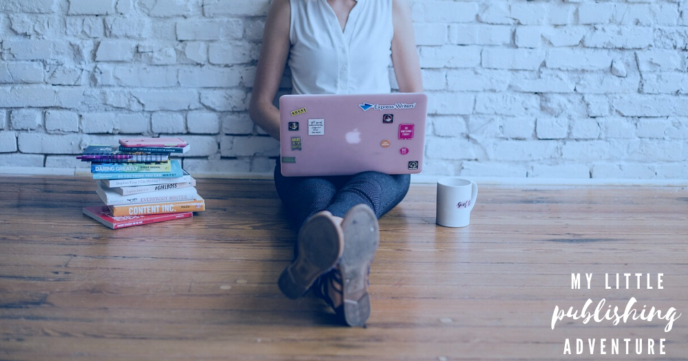 Girl sitting against a white brick wall, working on revisions on a pink laptop surrounded by books and coffee